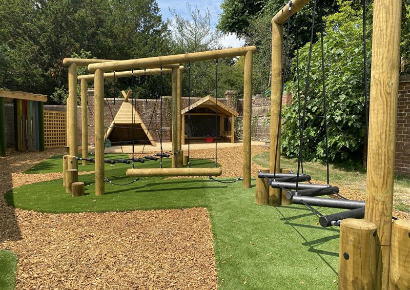 A wooden trim trail to the left of the photo with artificial grass placed below it. At the end is a wooden wigwam and playhouse, offering shelter from the sun. Wooden bark is seen surrounding the artificial grass.